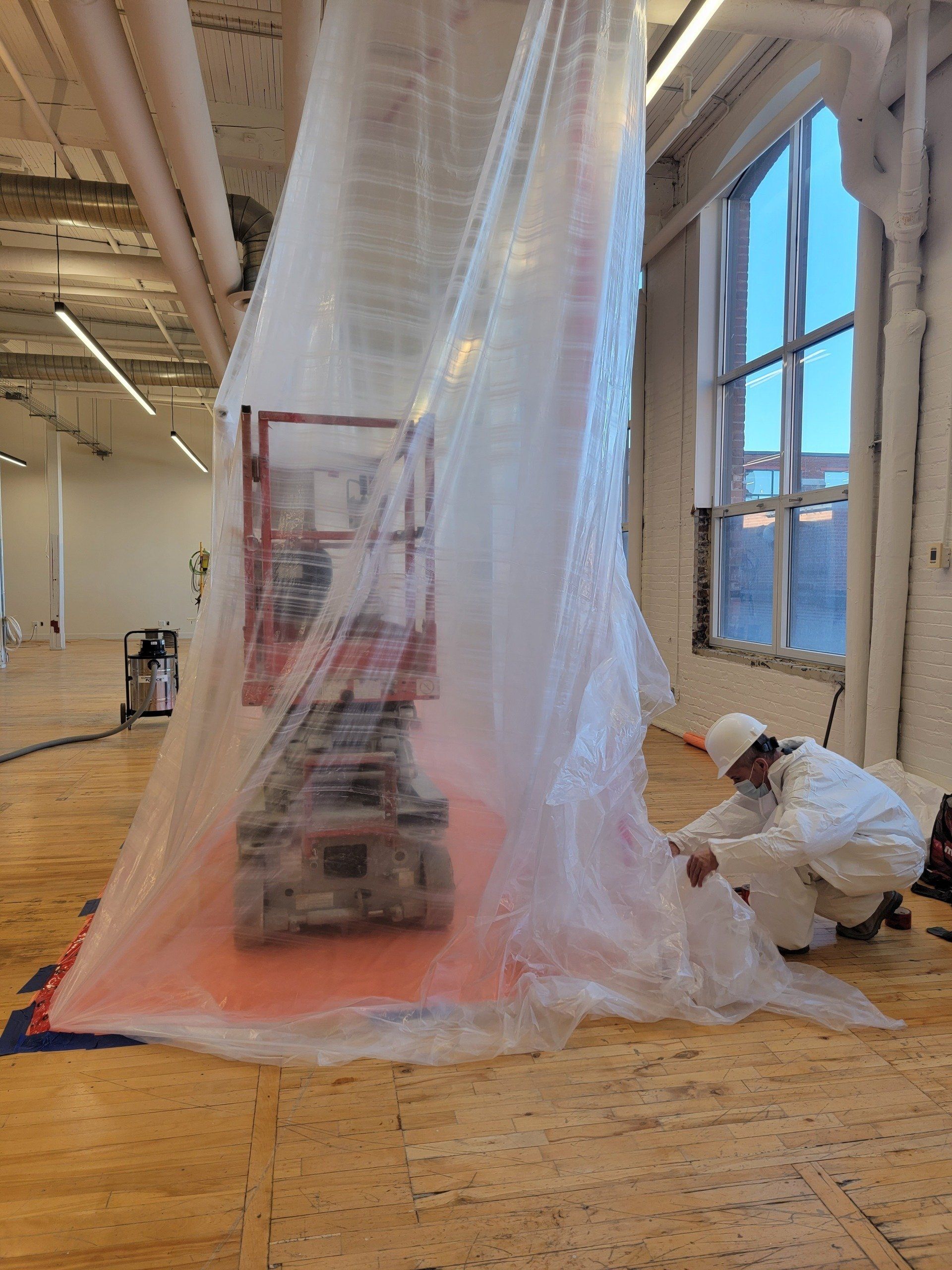 Ouvrier du bâtiment en combinaison de protection scellant un confinement en plastique autour de machines dans un bâtiment avec une grande fenêtre.