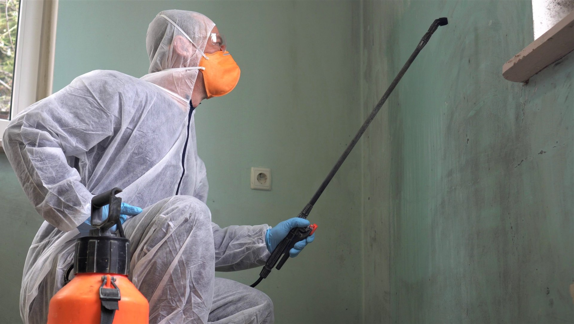 Une personne en combinaison de protection pulvérise un pesticide sur un mur. À l'intérieur, dans un coin d'une pièce.