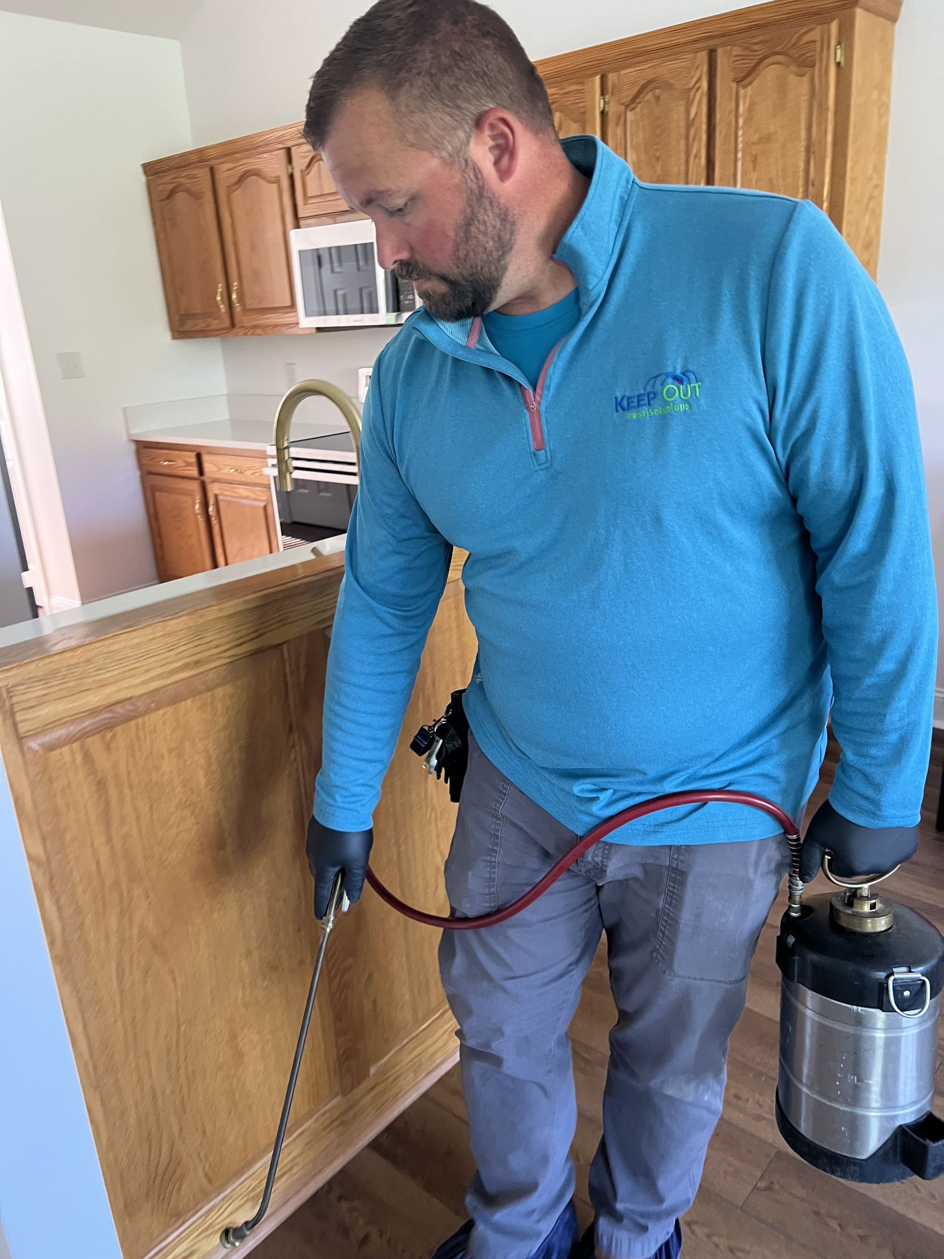 Matt Keep, owner of Keep Out Pest, in a blue shirt inspecting a wall in a kitchen, holding a canister and hose setup.