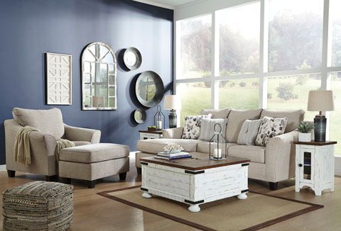 Living room with neutral-toned sofa and armchair, white and brown coffee table, white side table. Blue wall with decorative mirrors and natural light from the window.