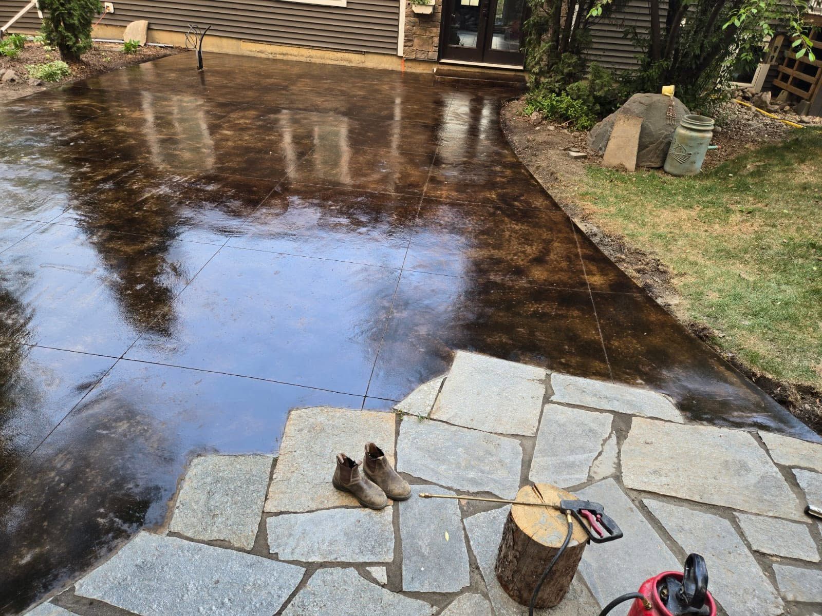 Wet stone patio with tools and a pair of shoes beside a freshly sealed driveway