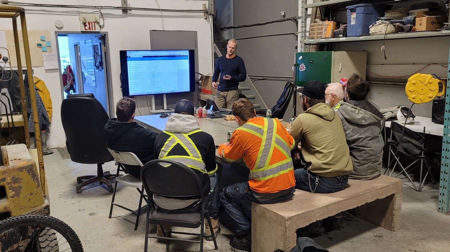 Workers in safety vests attend a training session in a workshop, watching a presentation on a screen.