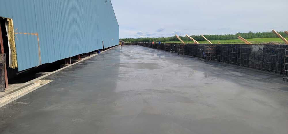 Wet concrete path beside a long blue building and stacked materials on a cloudy day
