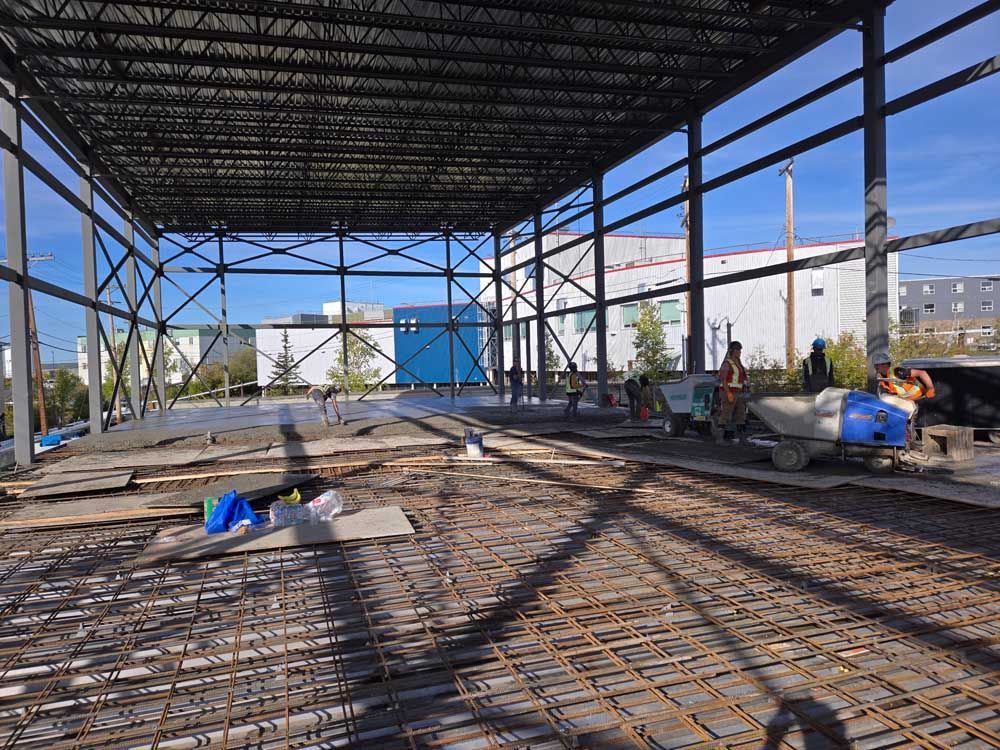 Interior of a steel-framed construction site with rebar floor, workers, and blue sky visible through the open walls