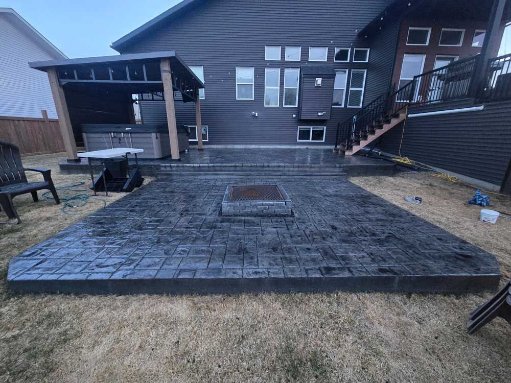 Backyard patio with a raised square fire pit and dark stamped concrete, beside a modern house.