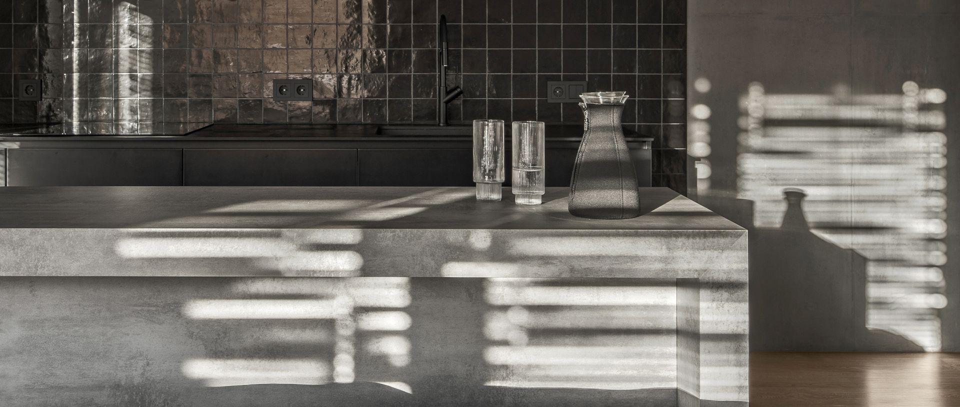 Modern kitchen counter with sunlit shadows, a tray, glasses, and a bottle on the counter