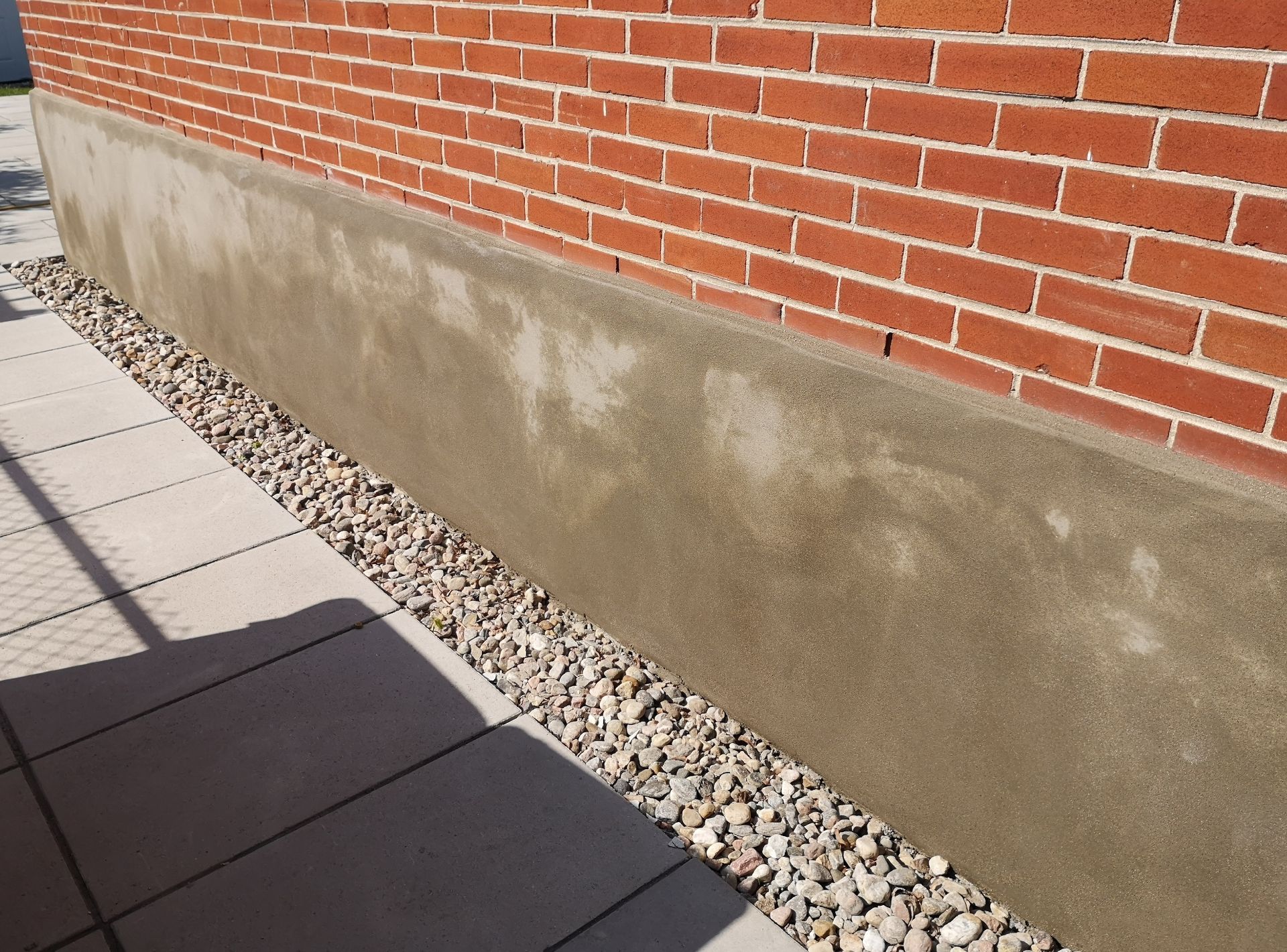 Brick wall with a weathered concrete base beside a gravel strip and paved walkway.