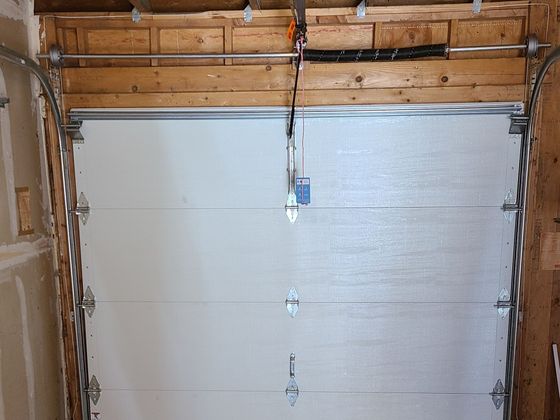 Inside a garage, a closed silver garage door with a hanging pull cord and exposed wood framing above.