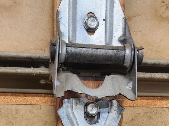 Broken metal hinge mounted on a tan door or panel, with visible screws and bent bracket.