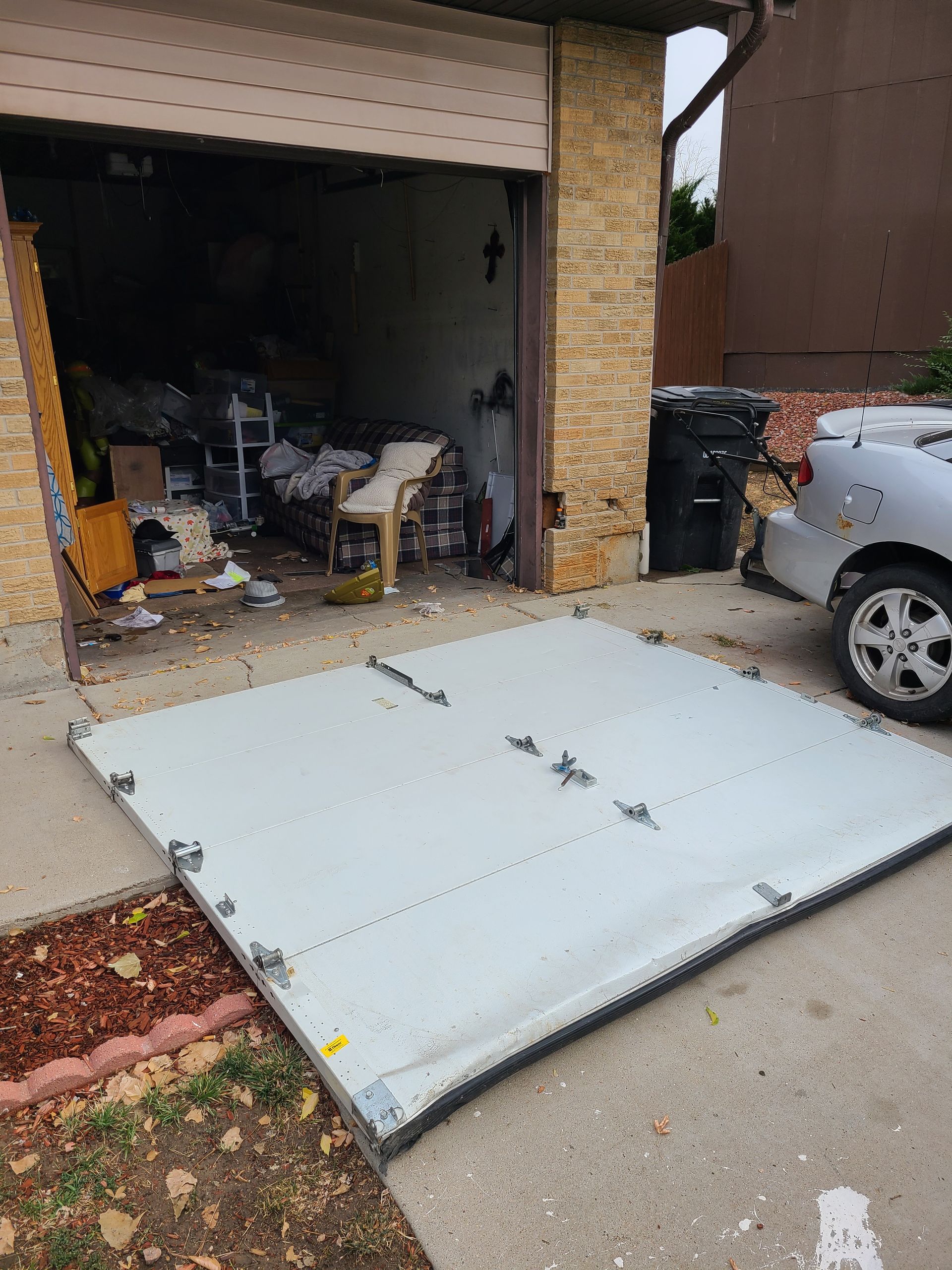 Collapsed garage door lying on driveway, with cluttered garage interior and parked car beside it.