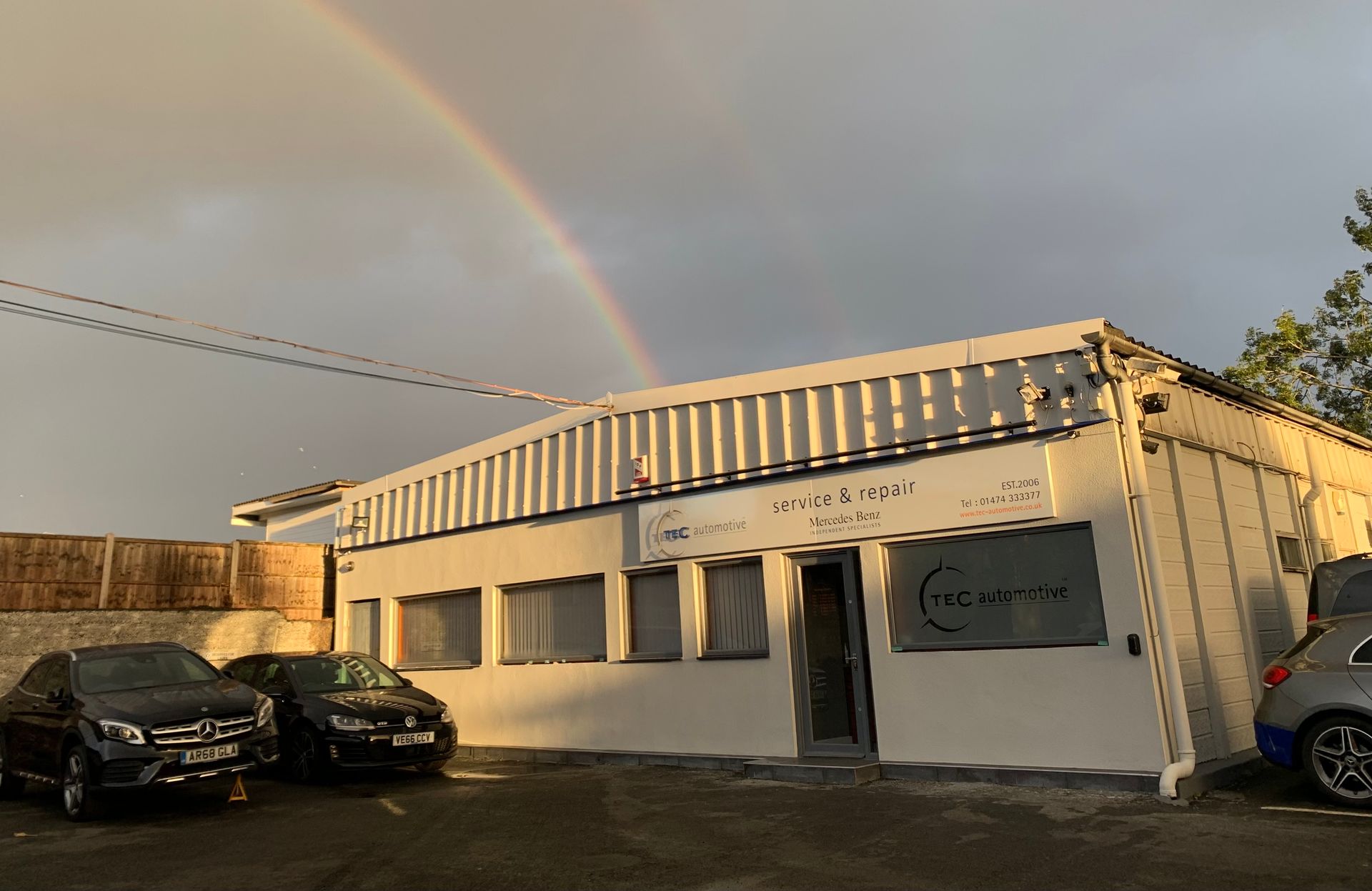A white building with cars parked in front of it and a rainbow in the sky.