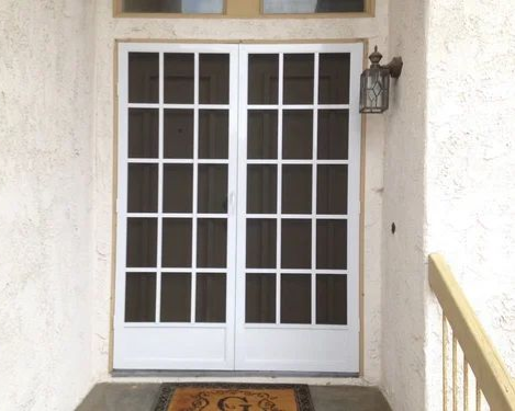 White double doors with multiple panes, flanked by stucco walls. A welcome mat rests at the entrance.