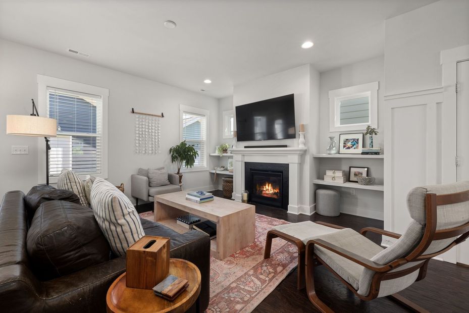 Modern living room with green sofa, fireplace, large wall TV, and wooden chairs near bright windows.