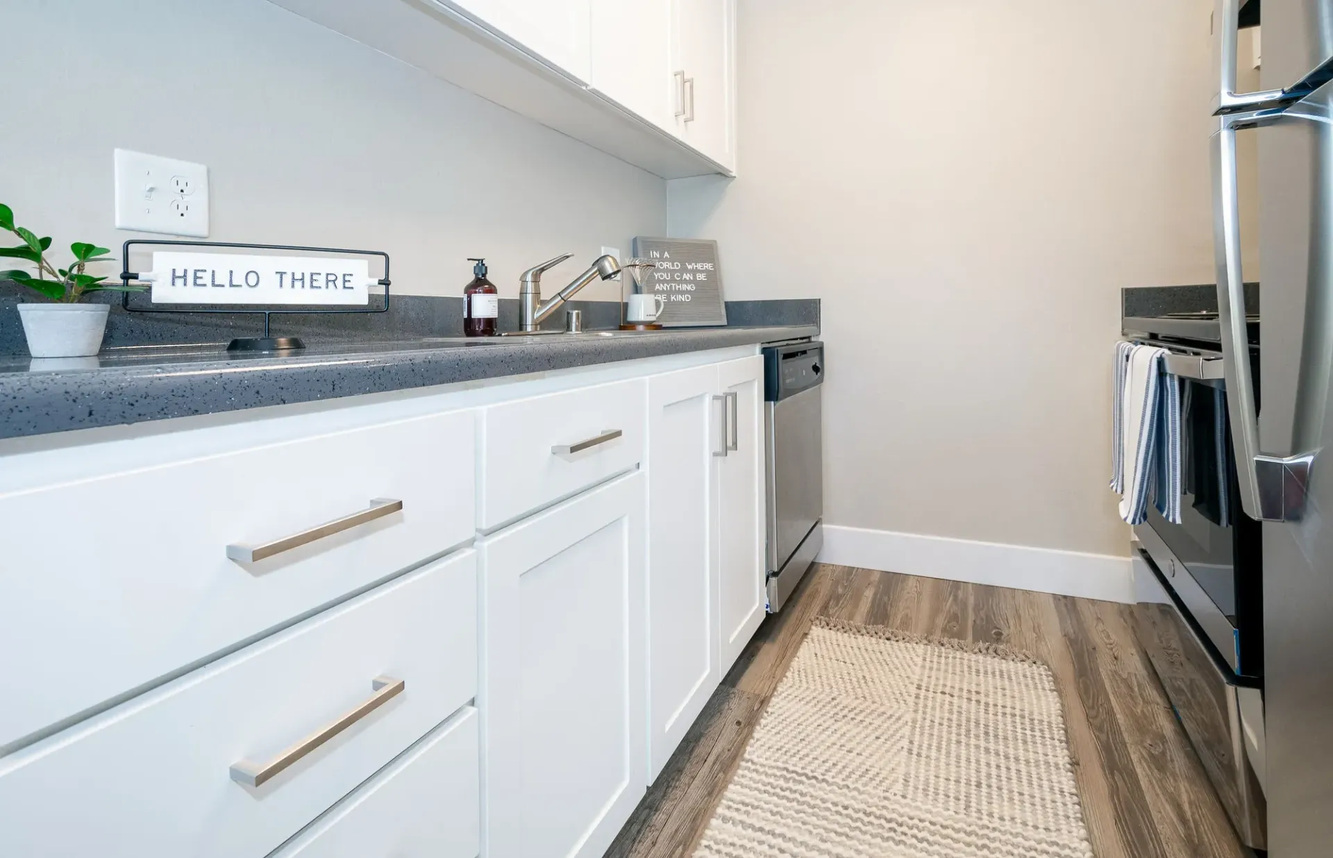 Bright kitchen with white cabinets, gray countertop, rug, and black stove along a narrow walkway.