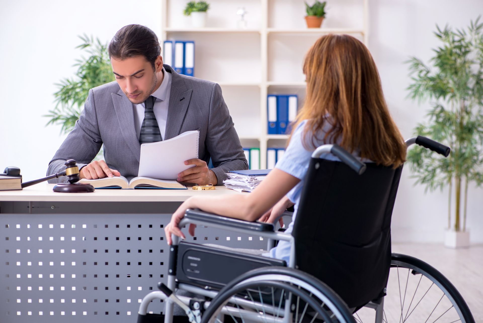 The backside of an injured woman in a wheelchair and a male lawyer in an office. Man in suit reviewing papers with woman in wheelchair at an office desk