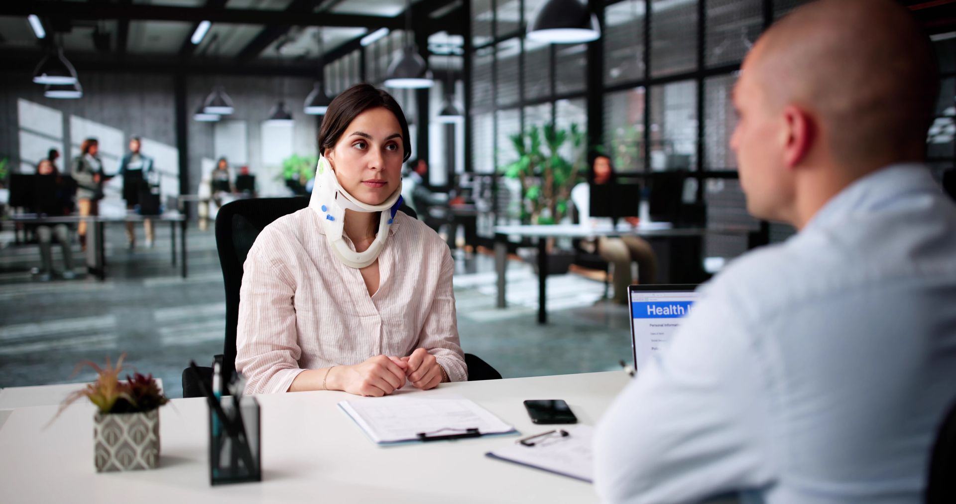 Two people in a modern office discuss across a desk, with papers, a laptop, and a small plant nearby.