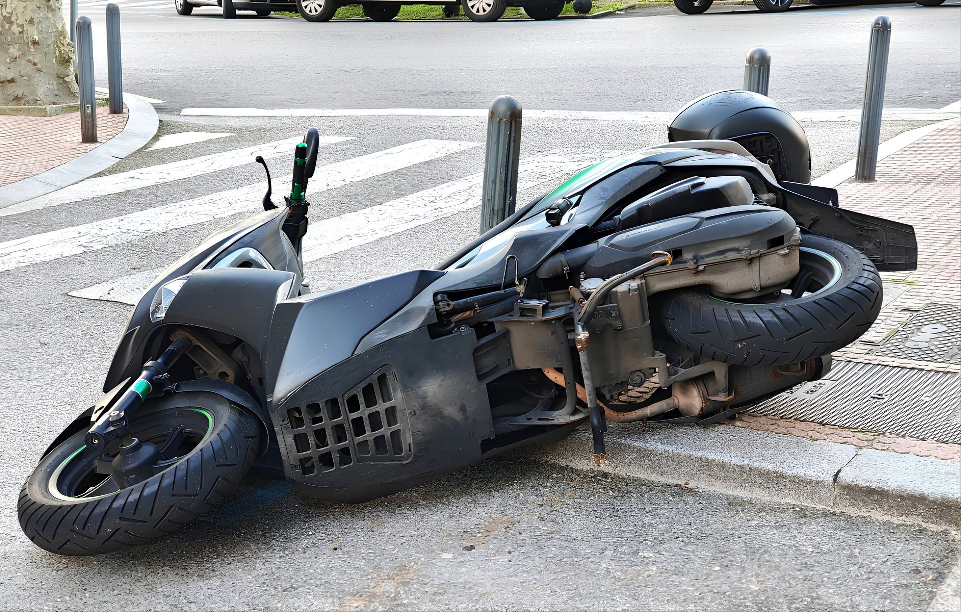Black scooter lying on its side on a curb in a street, with damaged bodywork and a detached rear wheel.