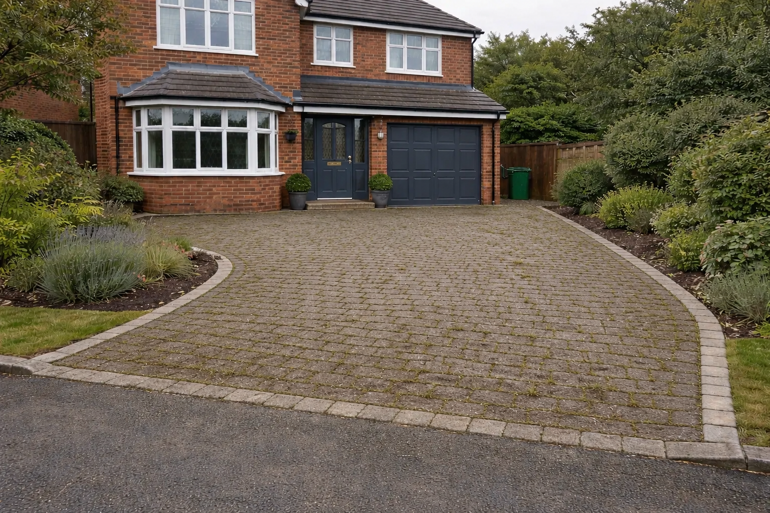 Curved asphalt driveway leading to a brick house with a black garage door and landscaped shrubs