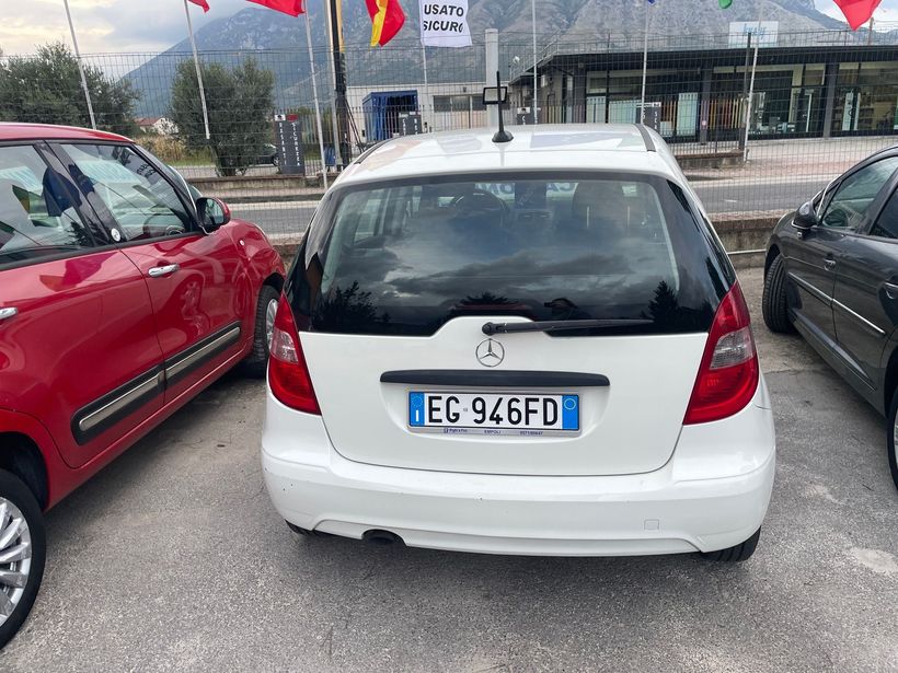 Vista posteriore di una Mercedes bianca parcheggiata tra auto rosse e scure in un parcheggio, targa visibile.