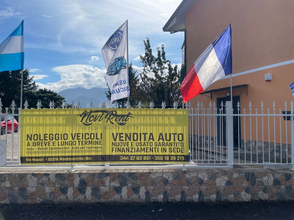 Concessionaria di auto con striscioni gialli per noleggio e vendita, bandiere e un edificio in mattoni sotto un cielo blu