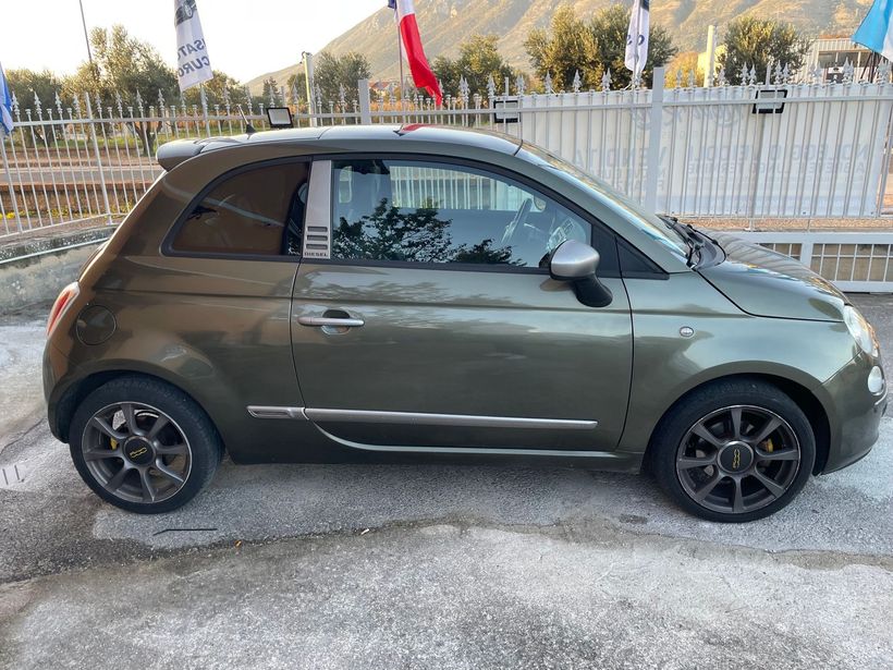 Una compatta hatchback verde scuro parcheggiata su una superficie ghiaiosa accanto a una recinzione e ad alcuni alberi.