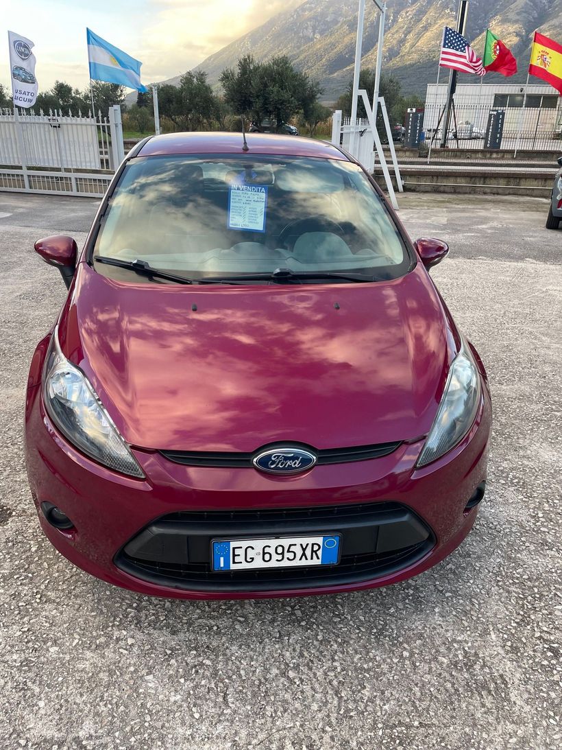 Una Ford hatchback rossa parcheggiata su un vialetto di ghiaia con montagne e bandiere sullo sfondo.
