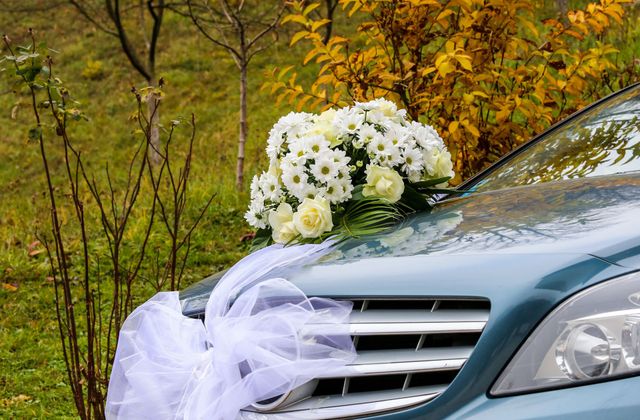 Un'auto argentata decorata con fiori bianchi e nastri di tulle, parcheggiata all'aperto tra le foglie autunnali.
