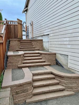 Stepped stone patio beside a white house, with brick retaining walls and railings up a sloped walkway