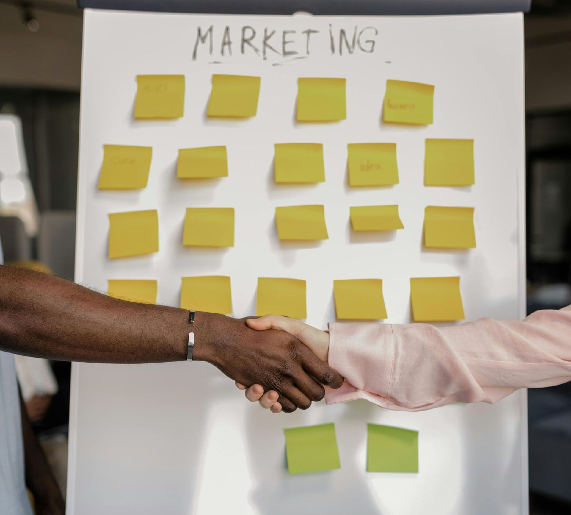 Close-up of two people shaking hands, with a blurred board in the background covered in sticky notes related to marketing strategies. Close-up of two people shaking hands, with a blurred board in the background covered in sticky notes related to marketing strategies.