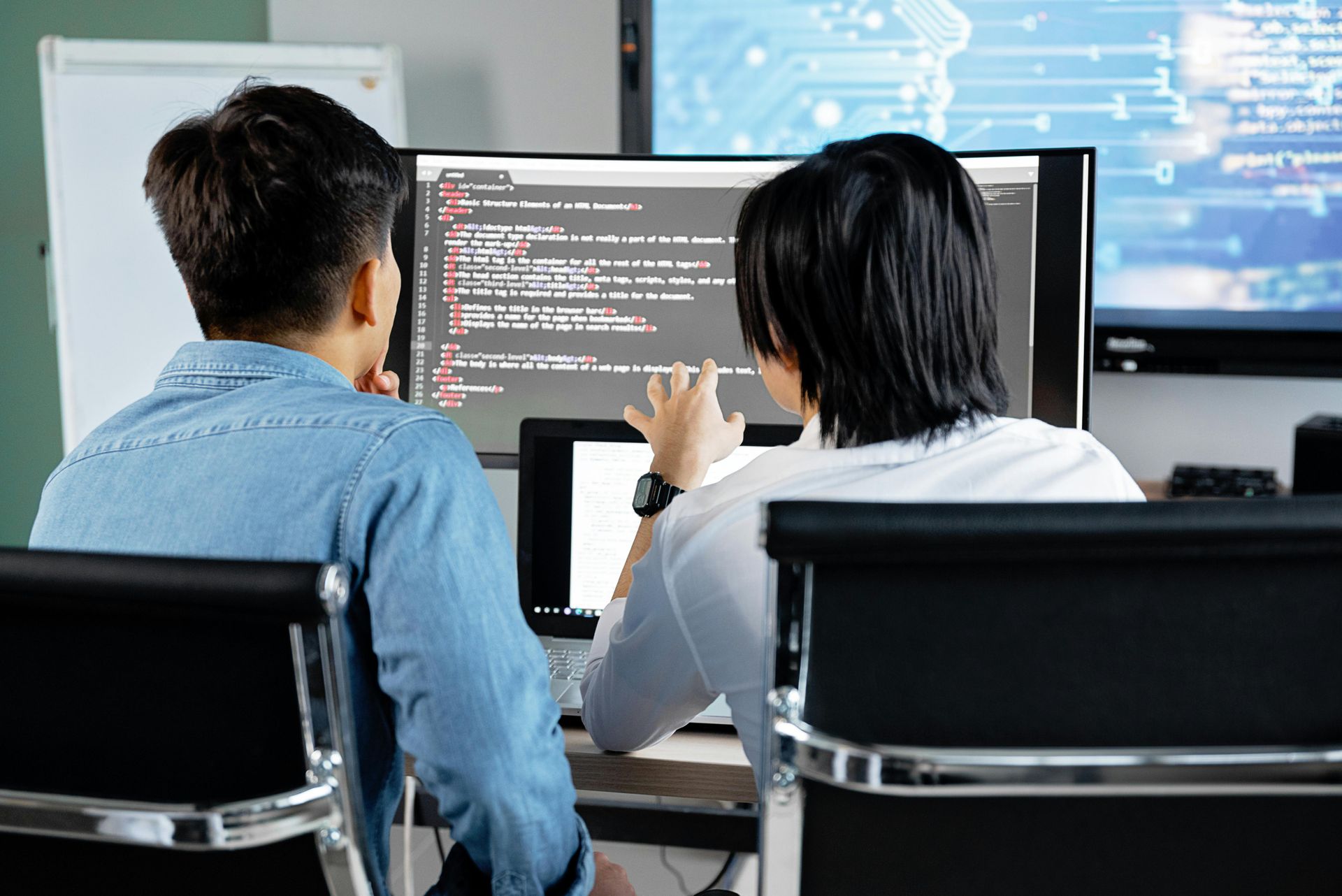 Two website developers sitting side by side in front of a monitor screen, engaged in a discussion while reviewing code or a web design project.