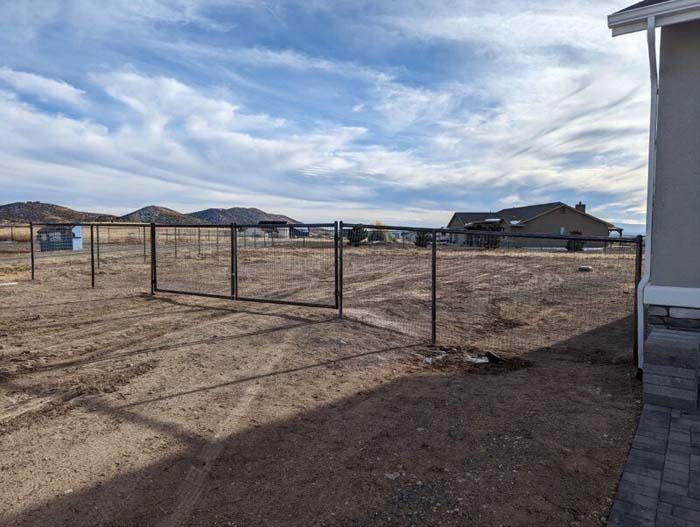 Empty fenced dirt yard beside a house under a cloudy sky, with mountains in the distance.