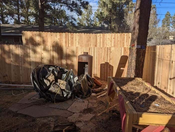 Backyard burn pile with charred debris beside a wooden fence and trees