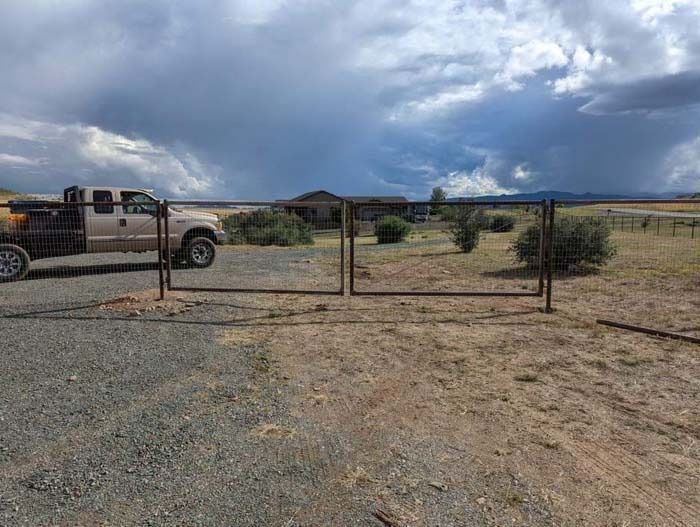 Rural dirt lot with a metal gate, parked pickup truck, and dark storm clouds overhead