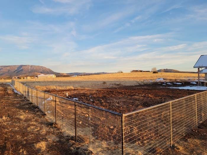 Fenced muddy field beside a road under a blue sky, with hills in the distance and a small structure on the right.