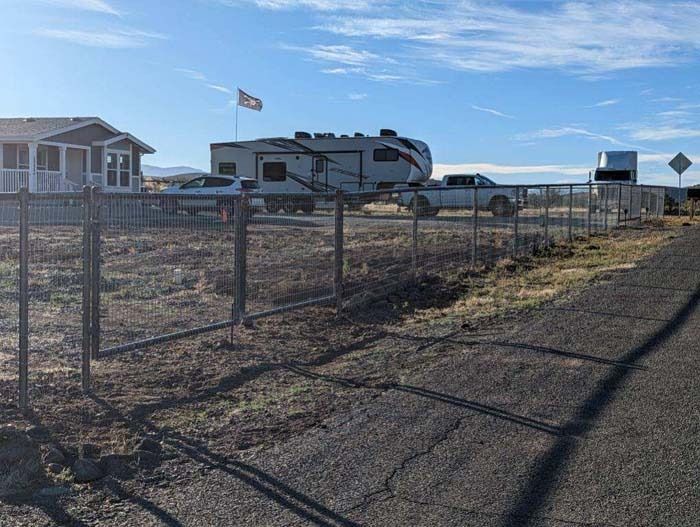 RV parked by a chain-link fence on a dusty roadside with a building and American flag in the background