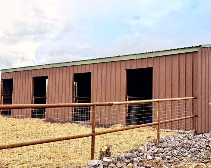 Long brown livestock barn with open stalls behind a metal fence and gravel foreground