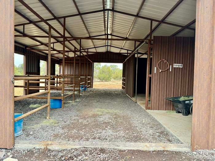 Open-sided barn interior with horse stalls, dirt floor, and a wheelbarrow near the back.