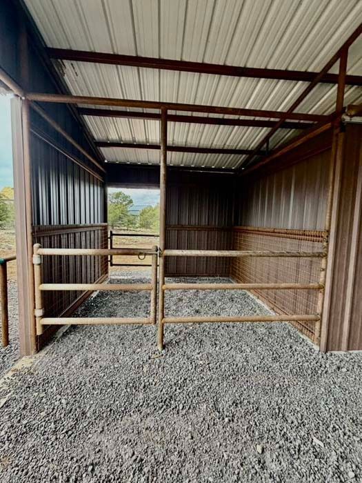 Empty livestock pen with gravel floor, metal rail fencing, and a corrugated roof opening to a grassy field.