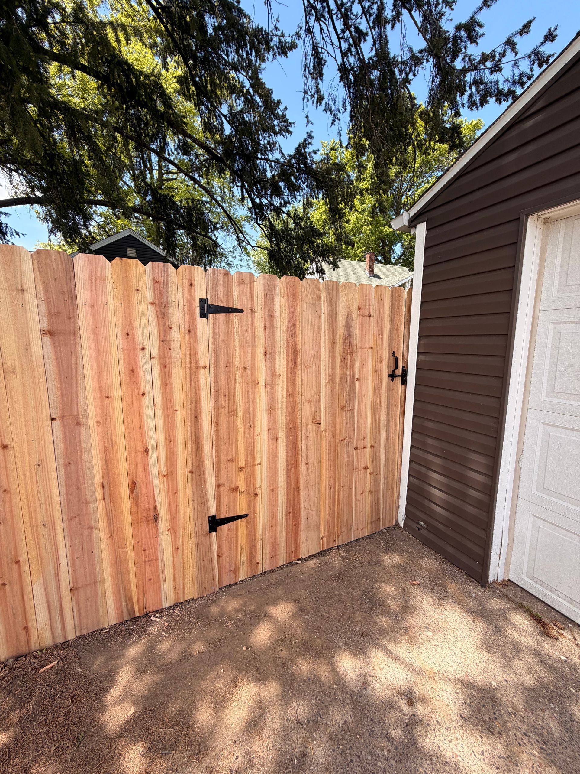 Wooden gate beside a brown garage, with trees casting shadows on the ground.