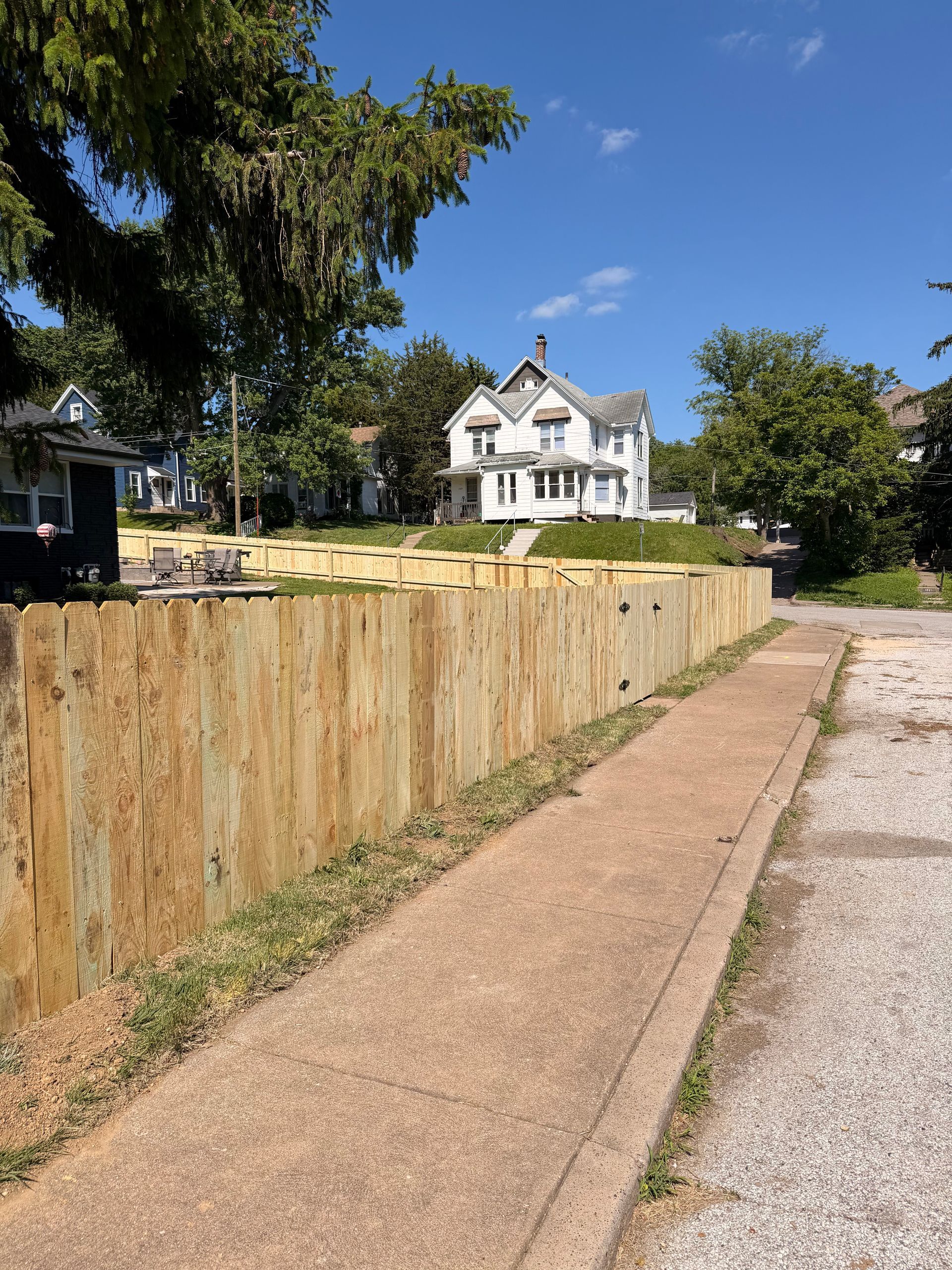 Residential sidewalk beside a wooden fence, leading toward a white house under a clear blue sky.