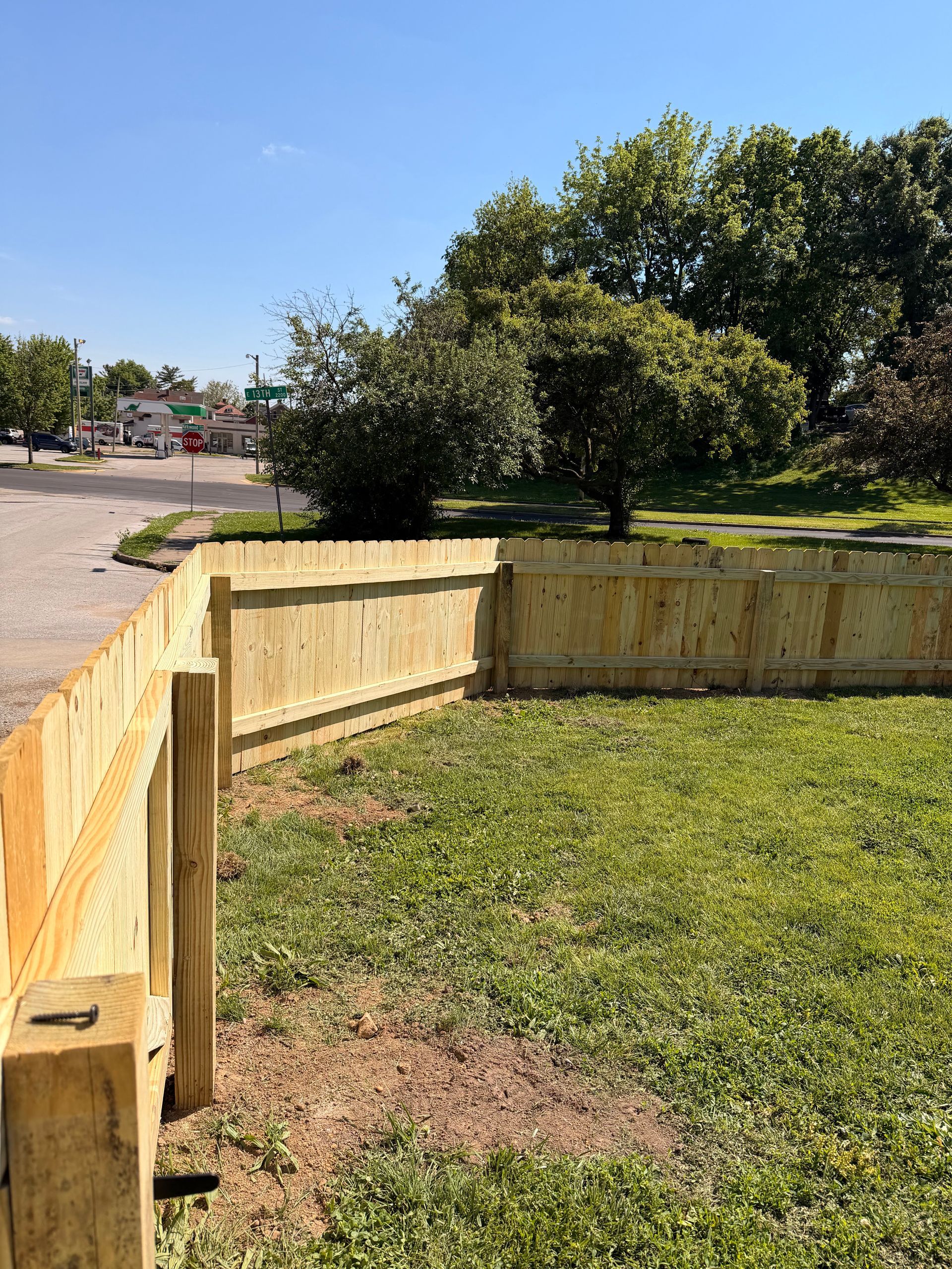 Wooden fenced backyard with green grass, trees, and a clear blue sky beside a gravel road.