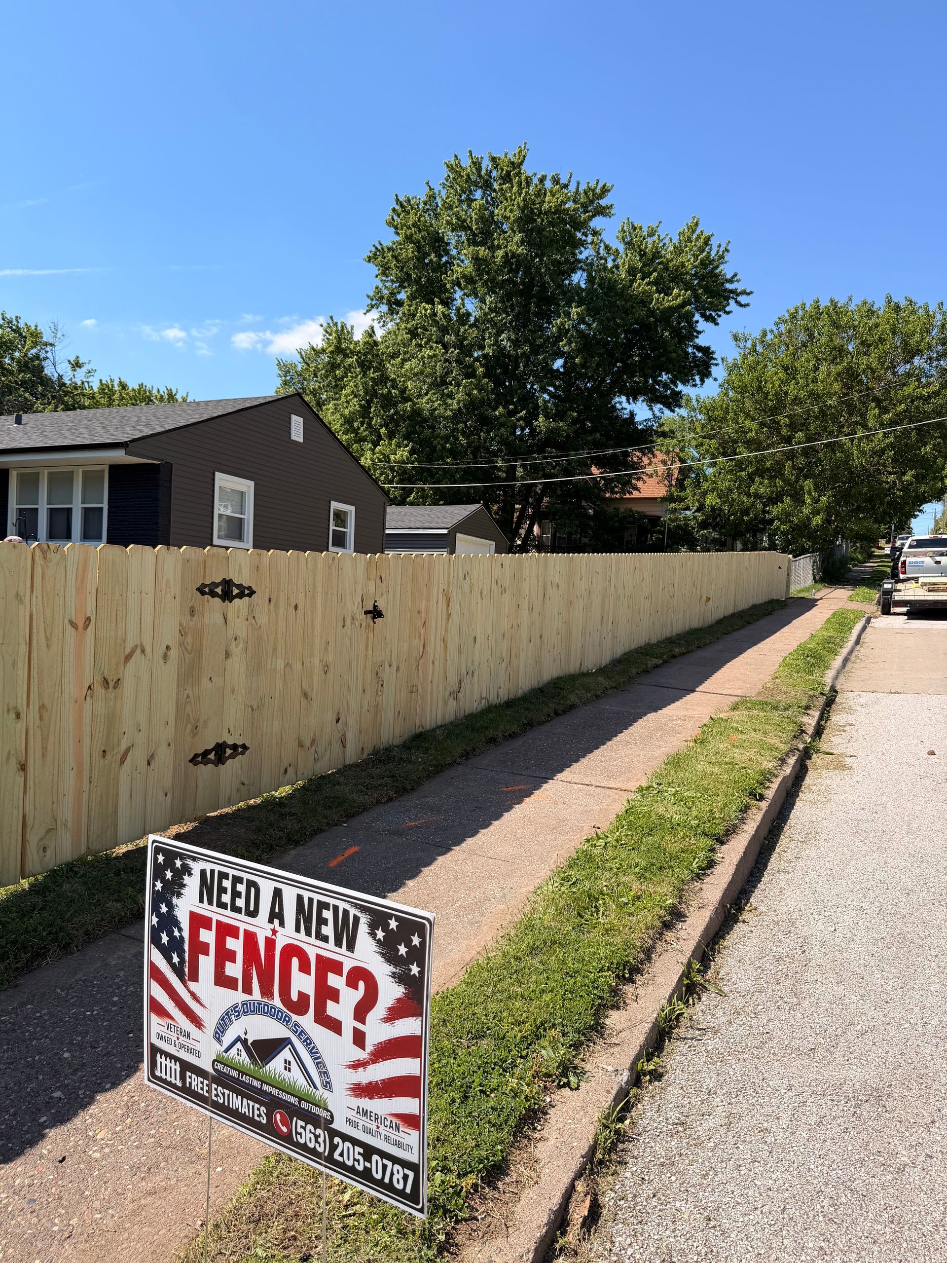 Wood fence along a gravel driveway with a “Need a Fence?” sign in the foreground under a sunny sky