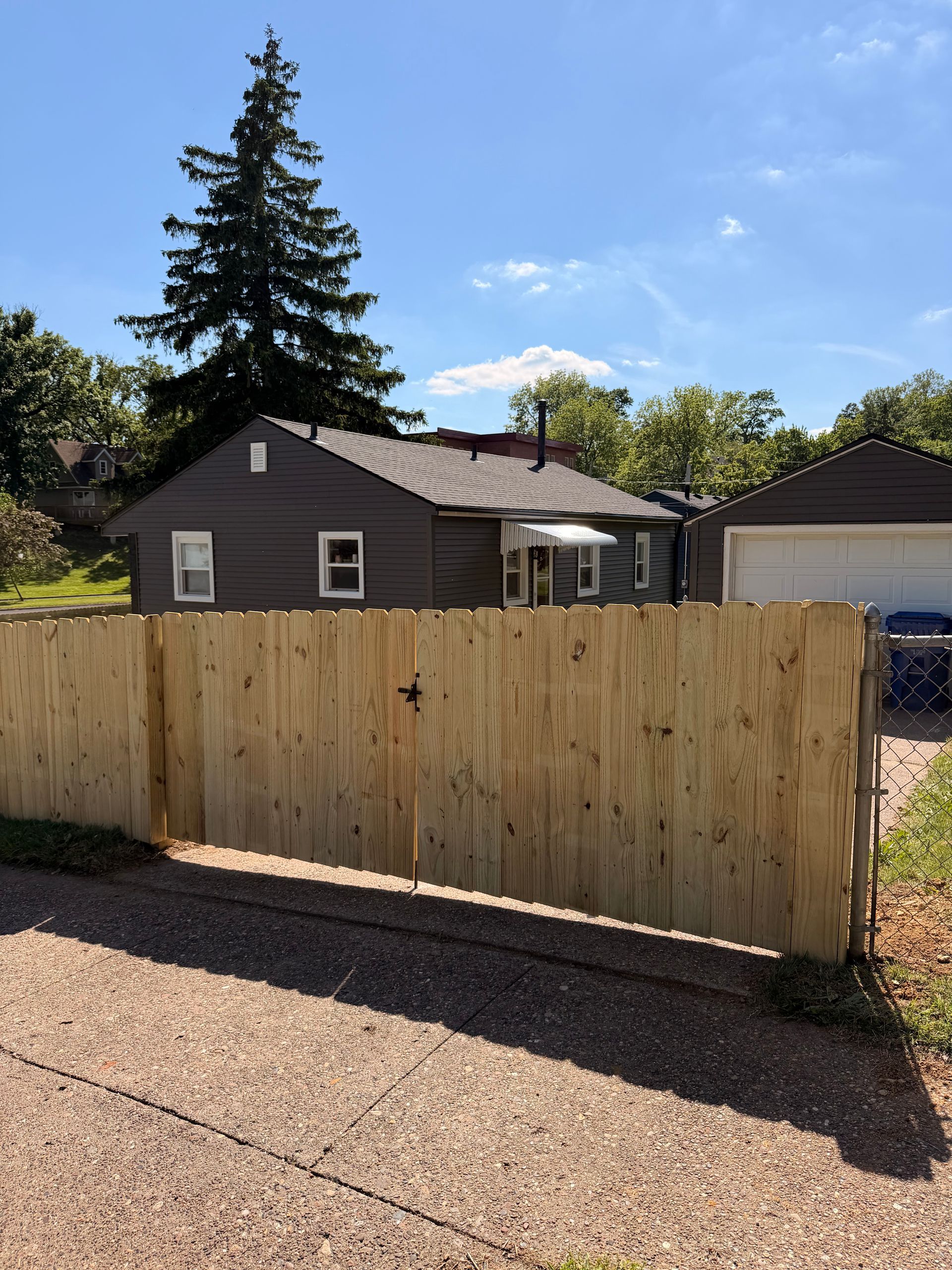 Wooden driveway gate in front of a gray house with a tall evergreen tree and blue sky