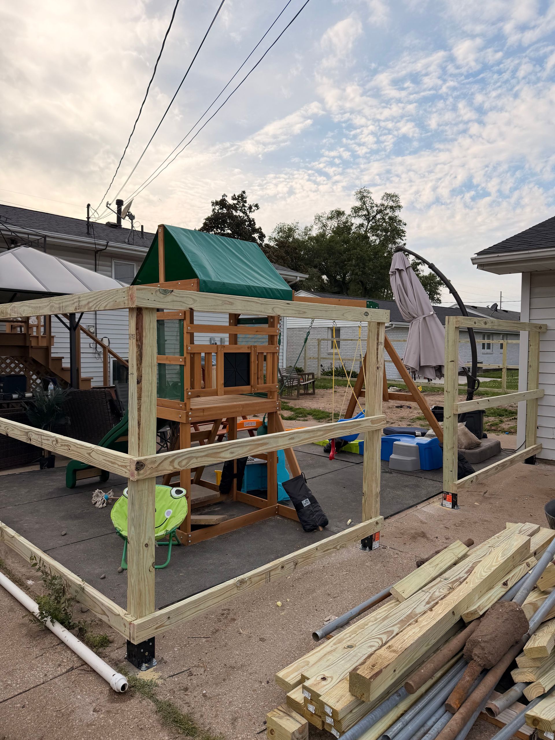 Wooden play structure under construction in a gravel yard, with tools, boards, and a green tarp roof