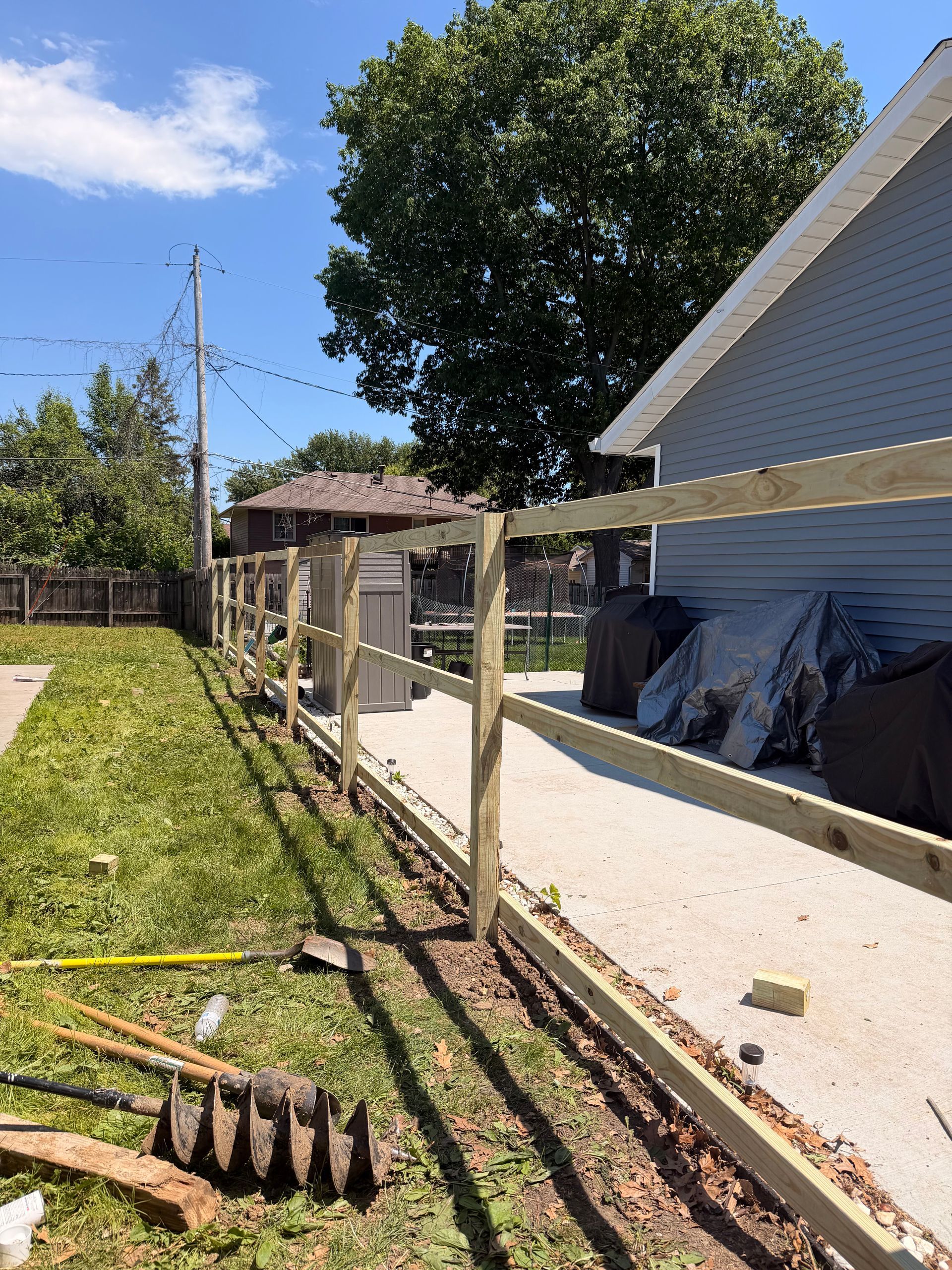 Backyard with fence posts, shed, lawn, and a house wall under a bright blue sky
