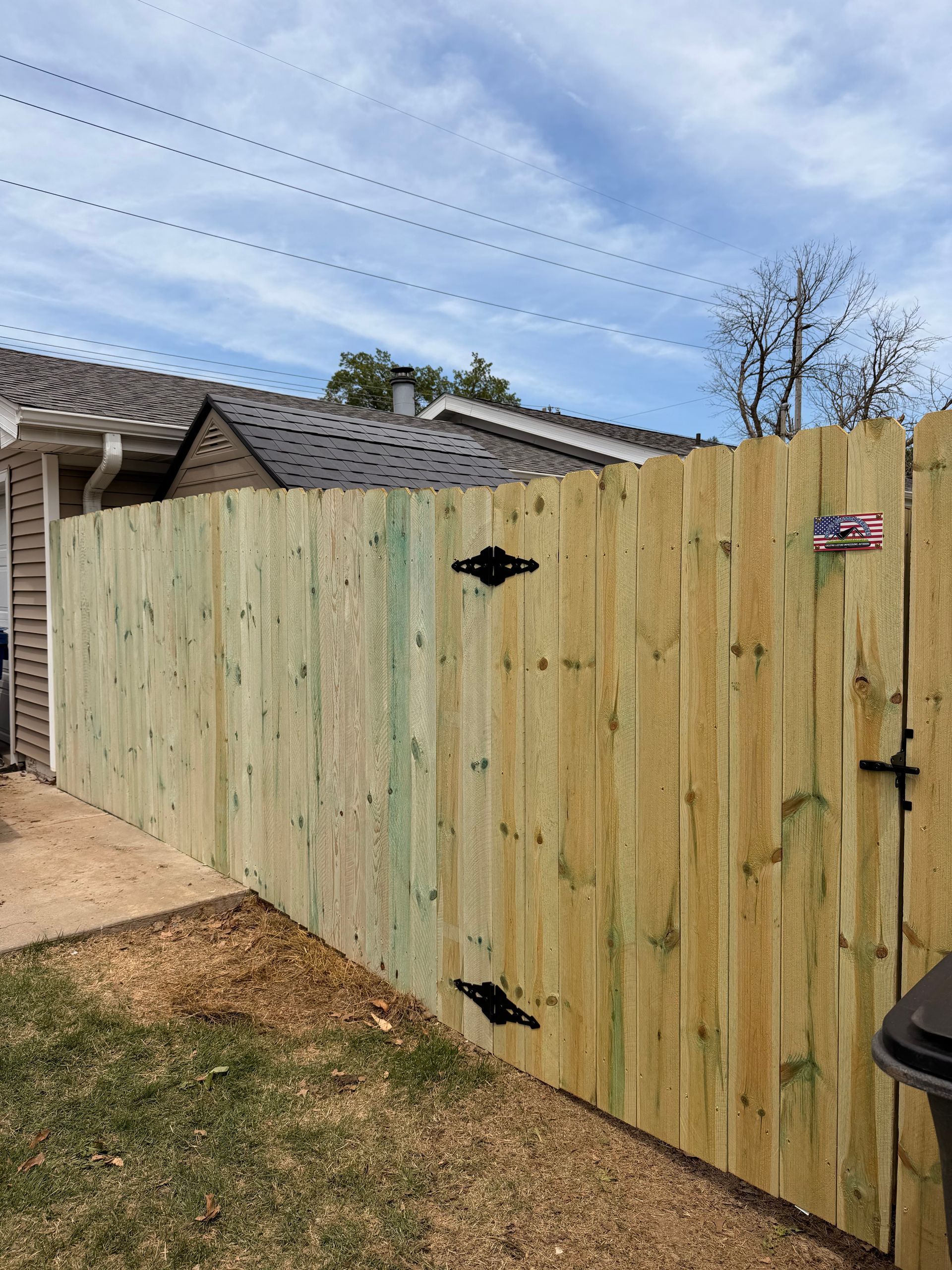 Wooden backyard gate with black hinges and latch beside a house under a blue sky