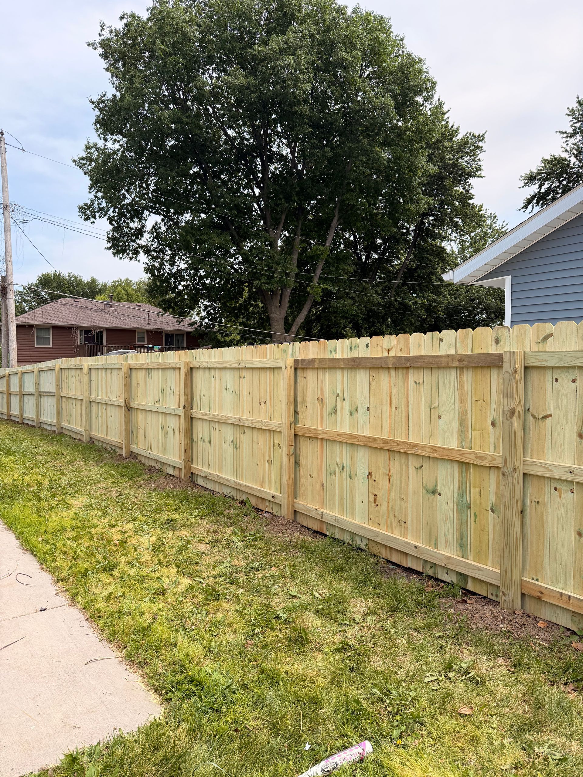 Wooden privacy fence along a sidewalk beside a grassy yard and a large tree under a clear sky.