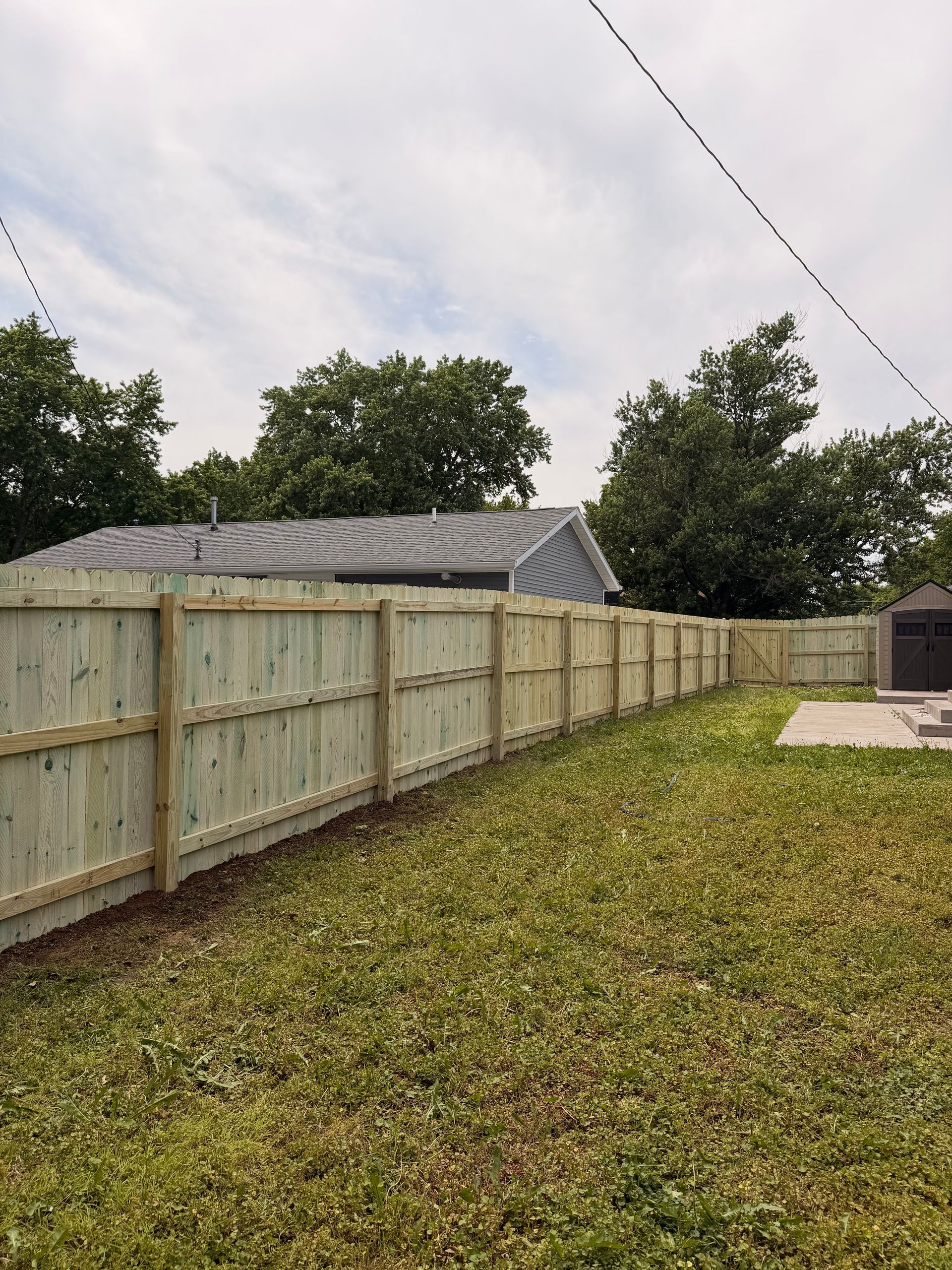Backyard with a long wooden fence, grass lawn, trees, and a small shed under cloudy sky