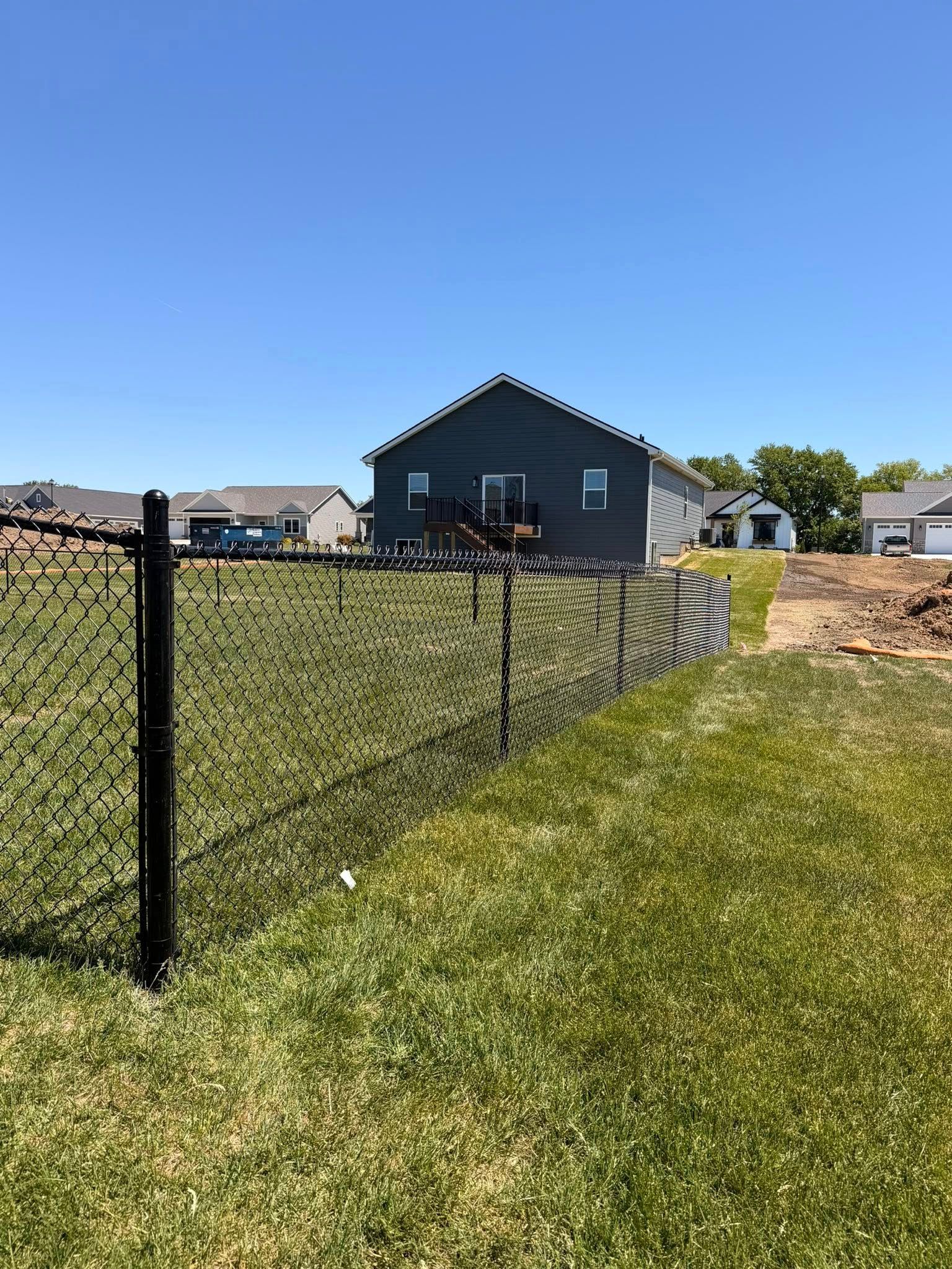 Black chain-link fence beside a grassy yard leading to a small gray house under a clear blue sky