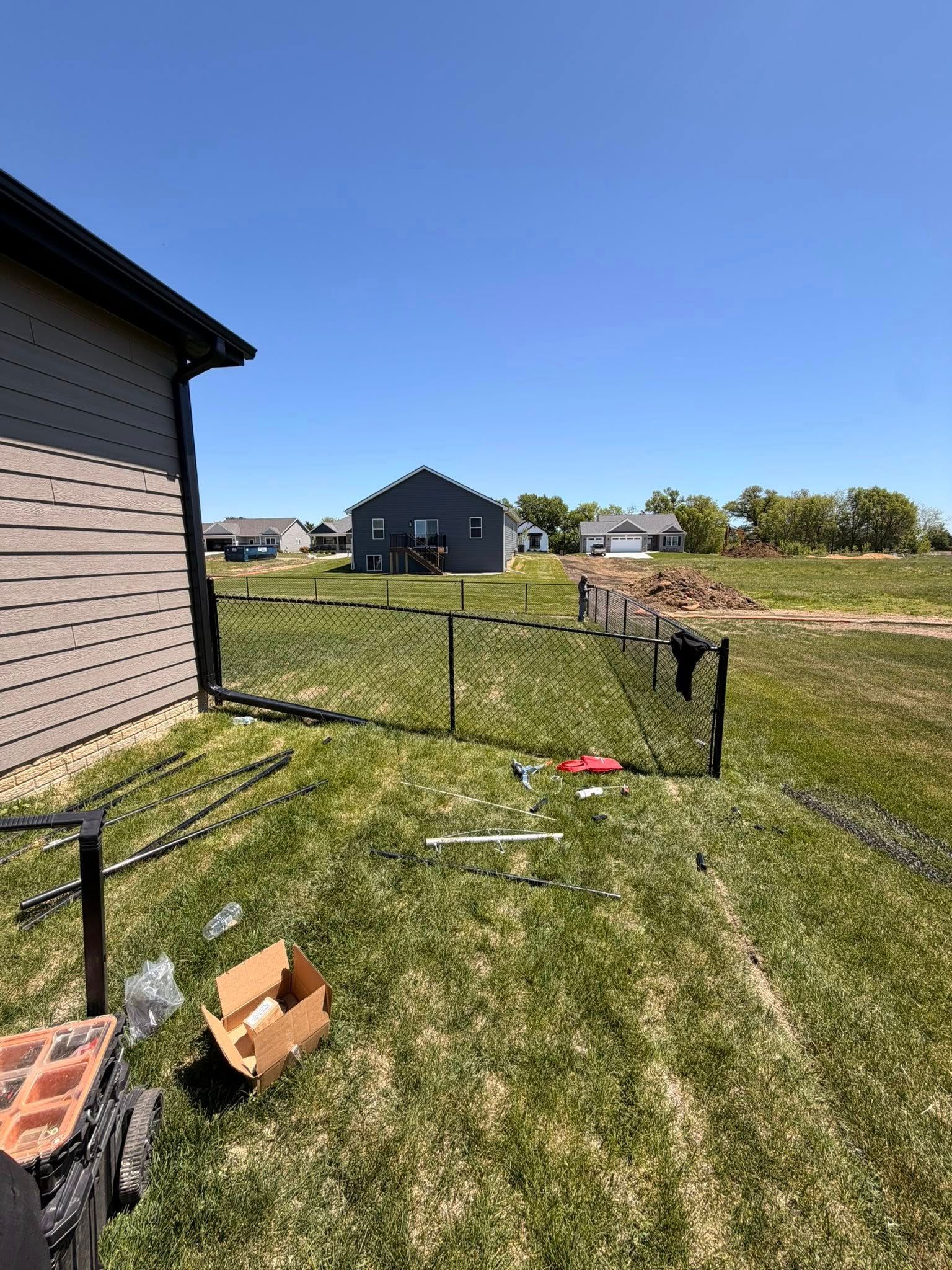 Backyard with a chain-link fence, a house on the left, and a small shed under a clear blue sky.